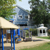 Blue/Brown tone children's playscape set in sand area with a white gazebo to the right and the blue/gray Fenton Township Hall
