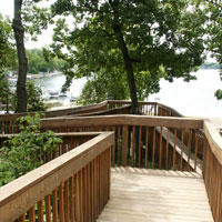 Brown stained wood boardwalk through the green trees with the water of the lake in the background