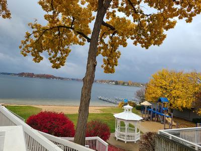 Gray sky mirrored on still water with a tree line of fall colors in the distance. There is the tan sand, red and yellow leaves.