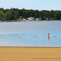 Photo foreground shows the sandy beach and the water line with orange swim area markers and the tree line in the background